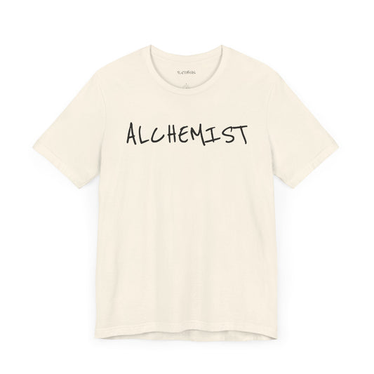 Natural t-shirt with handwritten black “ALCHEMIST” text across the chest